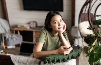 A young woman in a green t-shirt sits indoors, leaning on a cushion, holding a smartphone, and looking at the camera. A laptop and home decor are visible in the background.